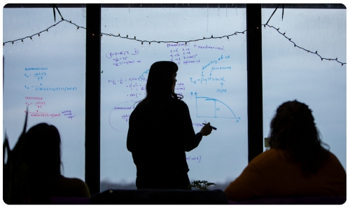 A faculty member in front of a glass board writing equations.